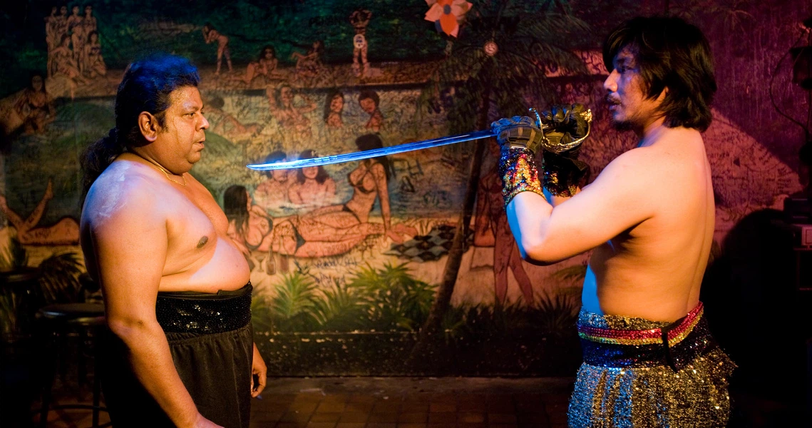 Two shirtless individuals are engaged in a performance, with one holding a sword pointed at the other's chest. They are standing in front of a colorful mural depicting various figures and scenes.