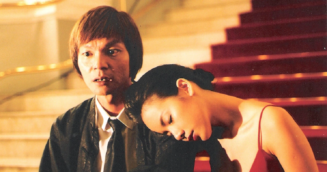 A film still from "One Leg Kicking" showing a young couple sitting on stairs, the woman resting her head on the man's shoulder.