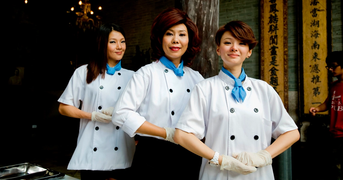 Three chefs in white uniforms stand together in front of a restaurant in a scene from the film Perfect Rivals.