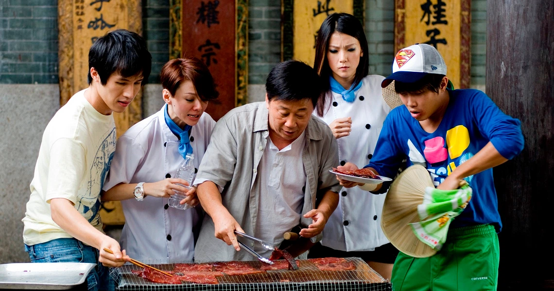 A scene from Perfect Rivals shows a group gathered around a grill, with chefs and others intently watching meat being cooked in a traditional setting.