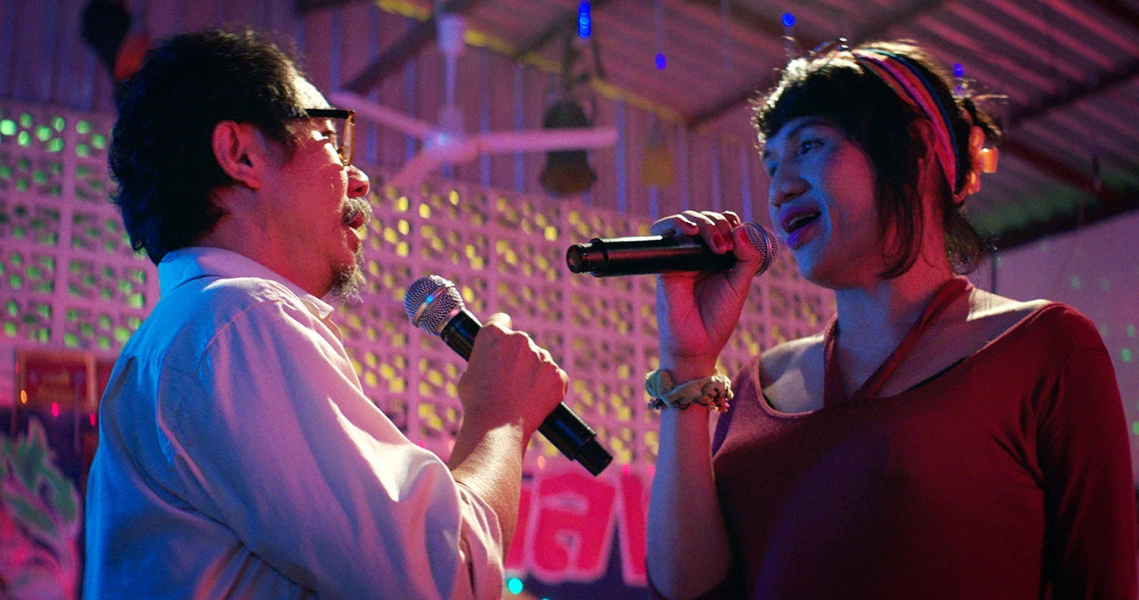 A scene from the film Pop Aye shows two characters singing karaoke together in a colorful, festive setting with neon lights.