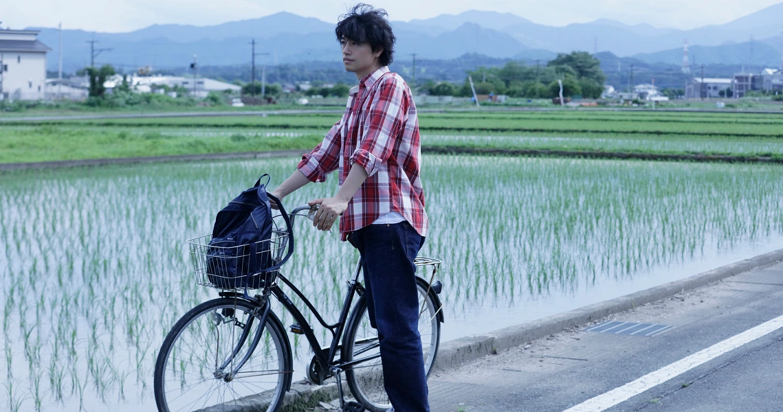 A scene from the film Ramen Teh shows a person with a bicycle standing on a road beside flooded rice paddies with mountains in the background.