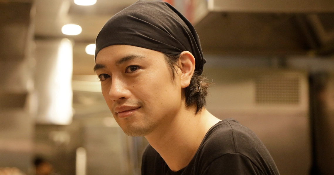 A close-up shot from the film Ramen Teh shows a young chef in a kitchen, wearing a black bandana and smiling warmly at the camera.