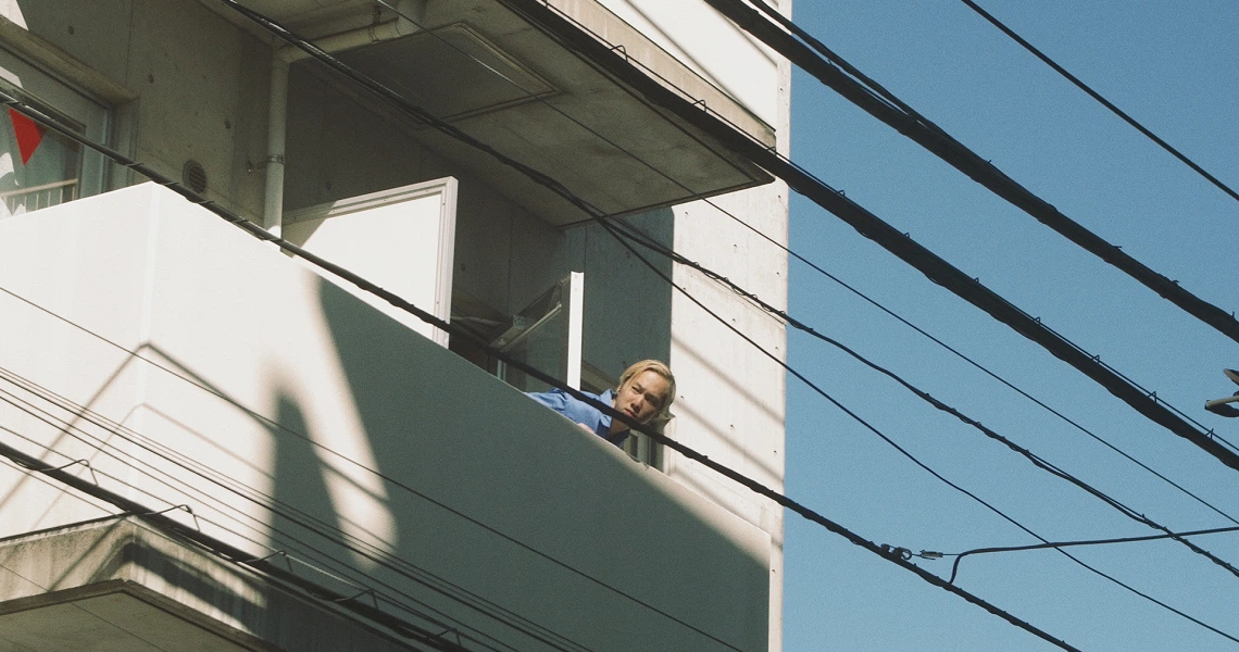 Revolution Launderette film still shows a person peering from an apartment balcony, with power lines in the foreground.