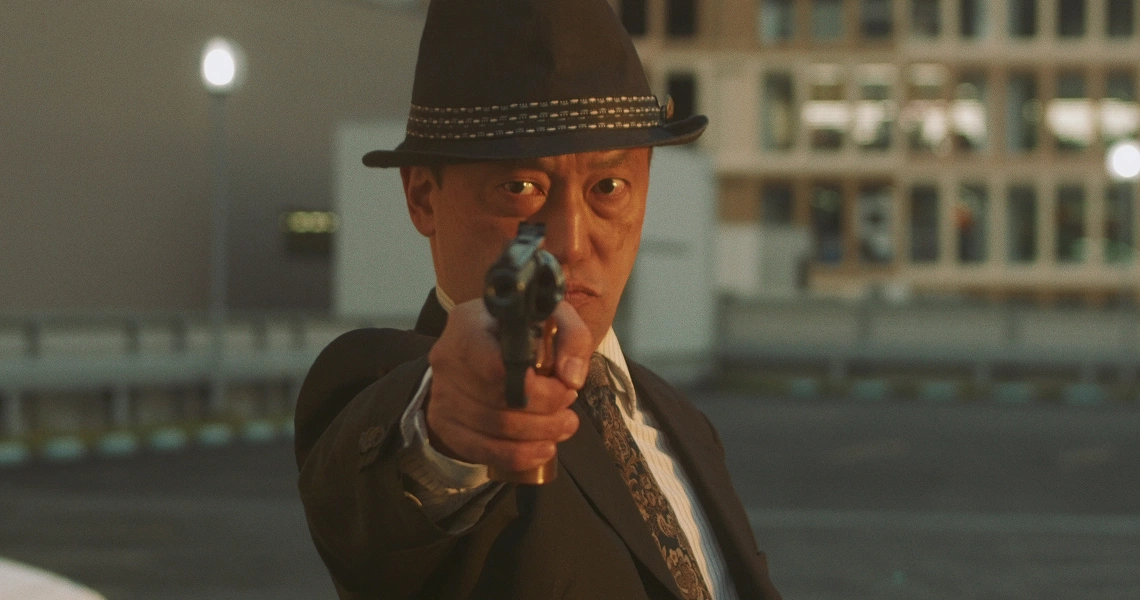 A tense scene from Revolution Launderette shows a man in a fedora aiming a gun directly at the camera, set against an urban backdrop.