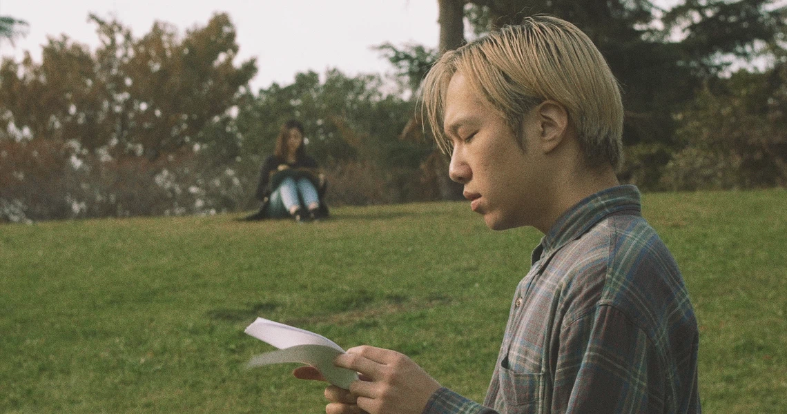 A scene from Revolution Launderette shows a young man reading in a park, with a woman sitting in the background.