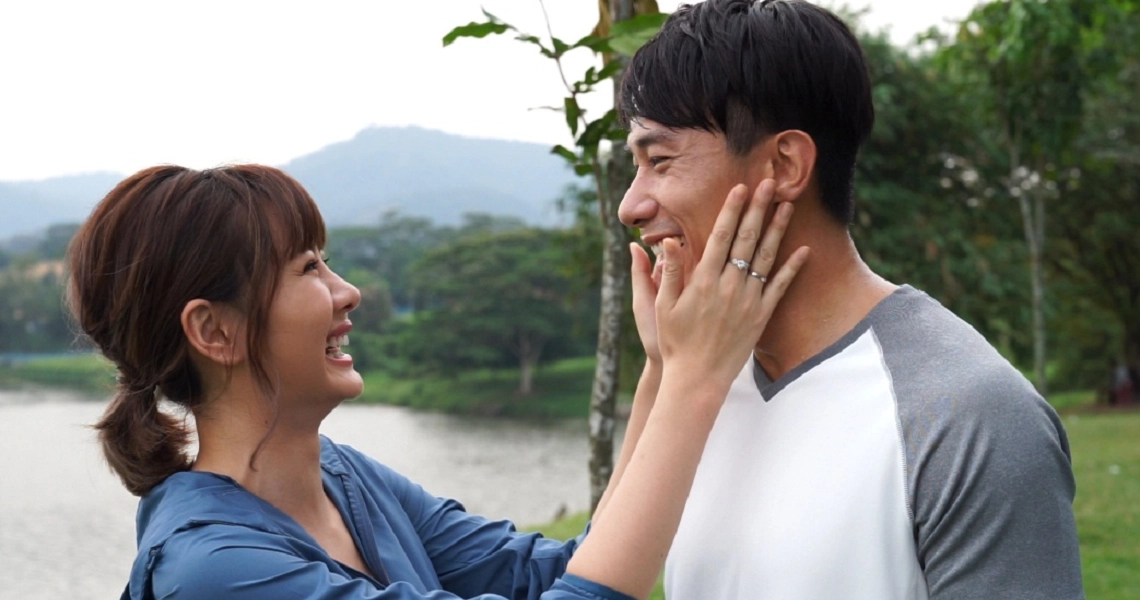 A romantic scene from "The Big Day" film, showing a couple embracing outdoors with a lake and mountains in the background.