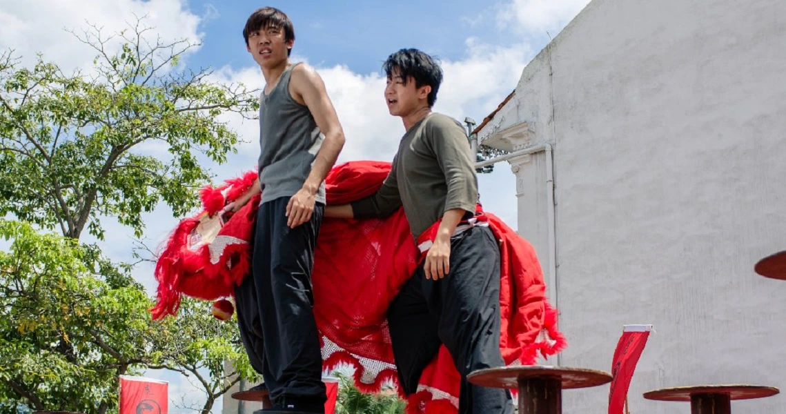 Two young men stand atop a vibrant red lion dance costume in a scene from the film "Lion Men," showcasing traditional Chinese culture.