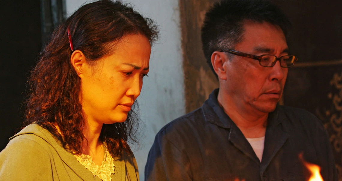 A scene from "The Maid" film showing two people standing solemnly in dim lighting, possibly during a candlelit vigil or ceremony.