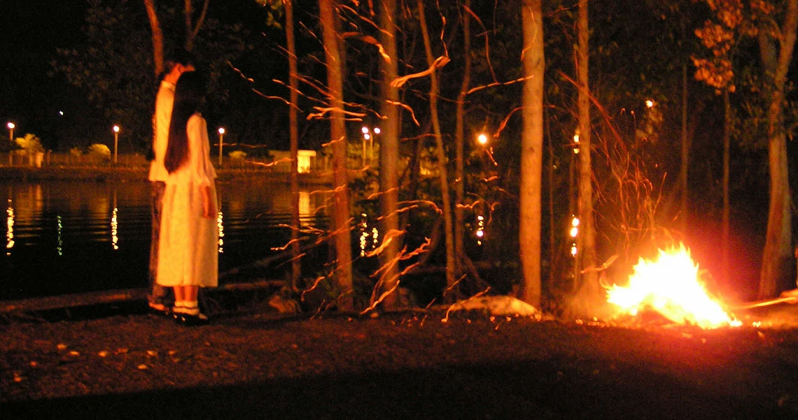 A haunting scene from "The Maid" film shows a figure in white standing near trees, with a fire burning in the foreground.