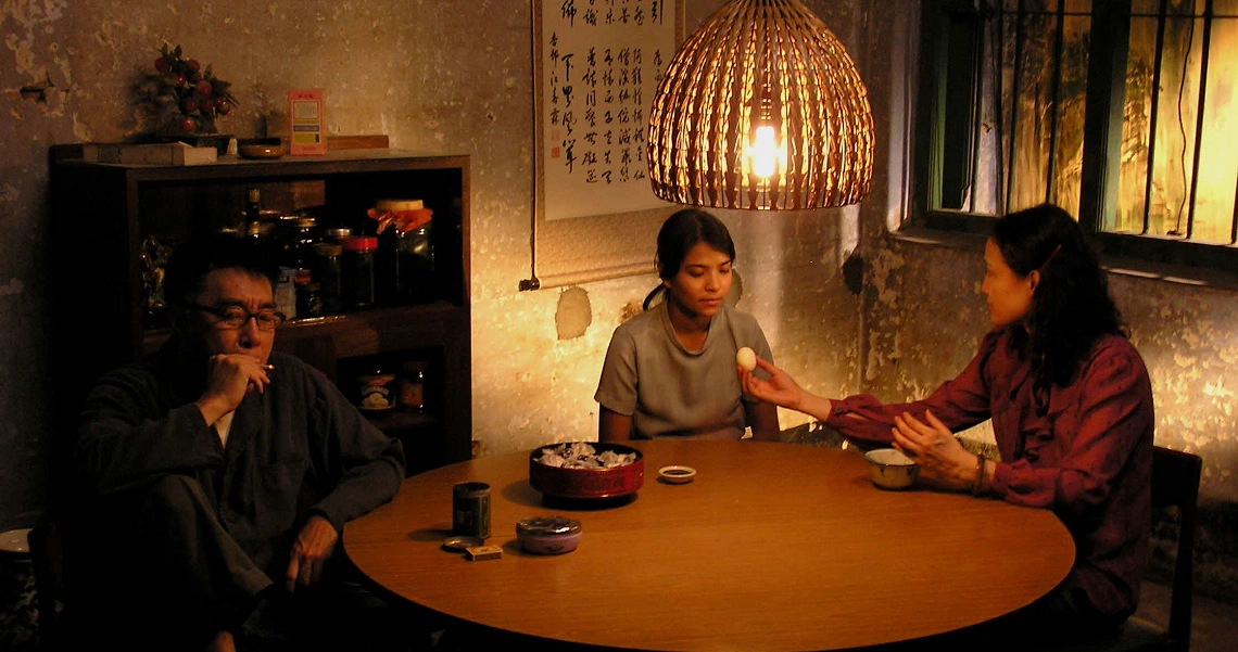 A dimly lit scene from "The Maid" film showing three people seated around a table in a cozy room with warm lighting.