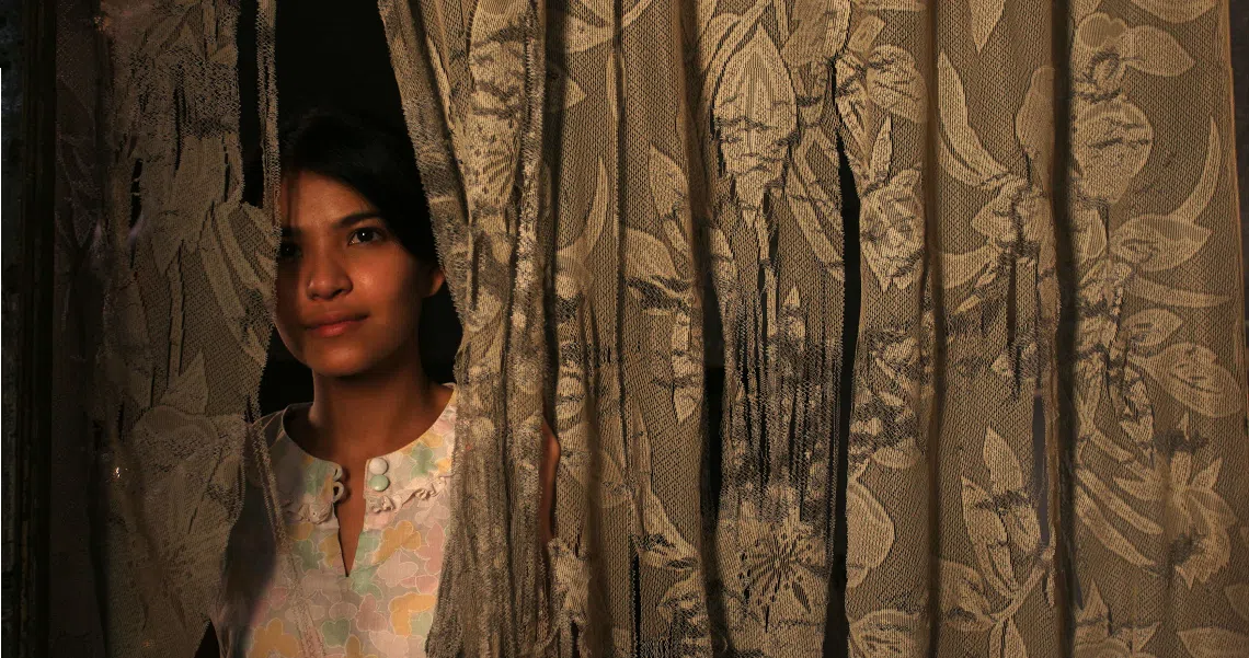 A young woman peeks through ornate curtains in a film still from "The Maid," creating an atmosphere of mystery and intrigue.