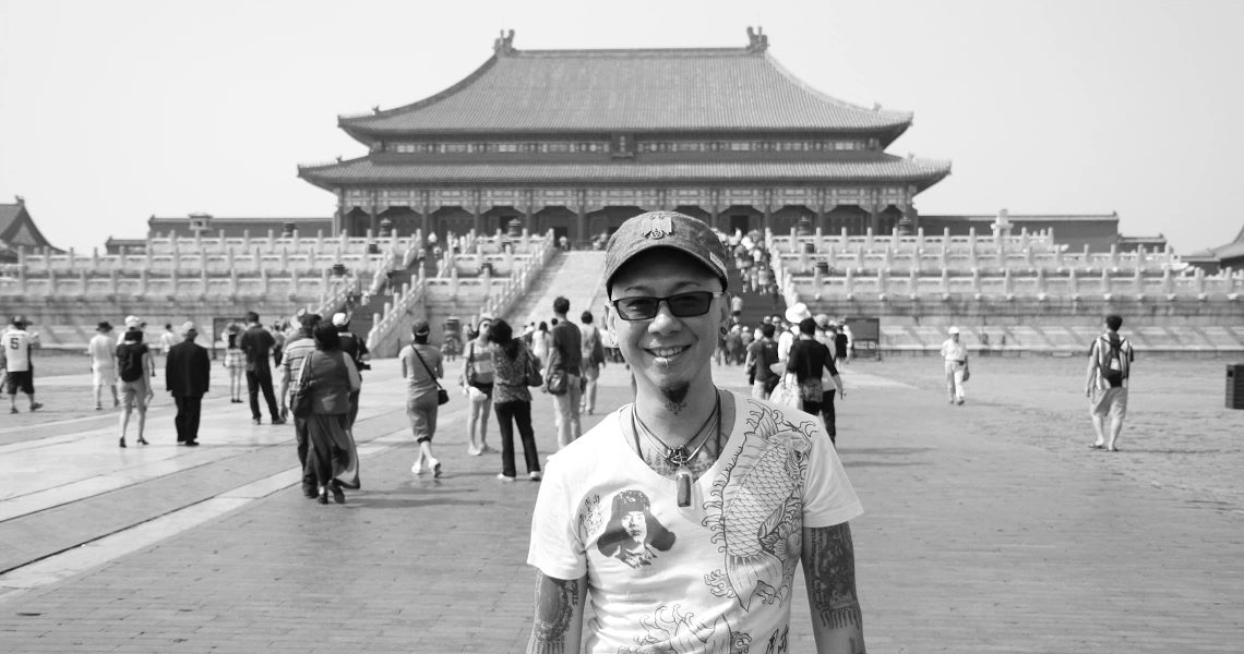 A film still from "The Naked DJ" showing a man in sunglasses and a t-shirt standing in front of a traditional Chinese building.