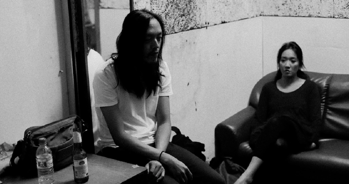 A black and white still from "The Obs: A Singapore Story" showing two women sitting in a room, one on a couch and one on the floor.