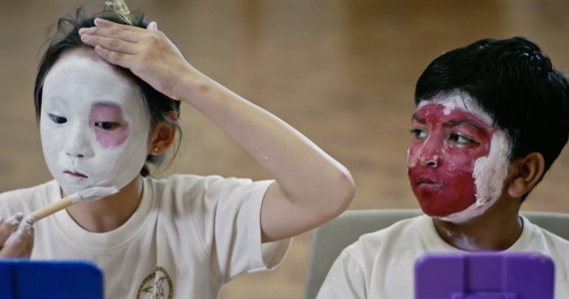 Two children with painted faces in a scene from The Wayang Kids film, one with white makeup and one with red.