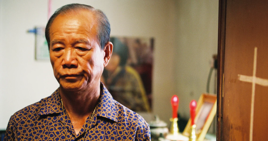 A still from the film "Truth Be Told" shows an elderly Asian man with a serious expression, wearing a patterned shirt in an interior setting.