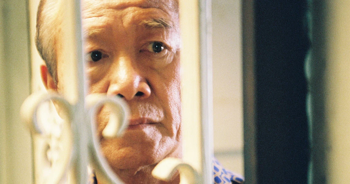 A close-up still from the film "Truth Be Told" shows an elderly man peering through a window or doorway with a concerned expression.