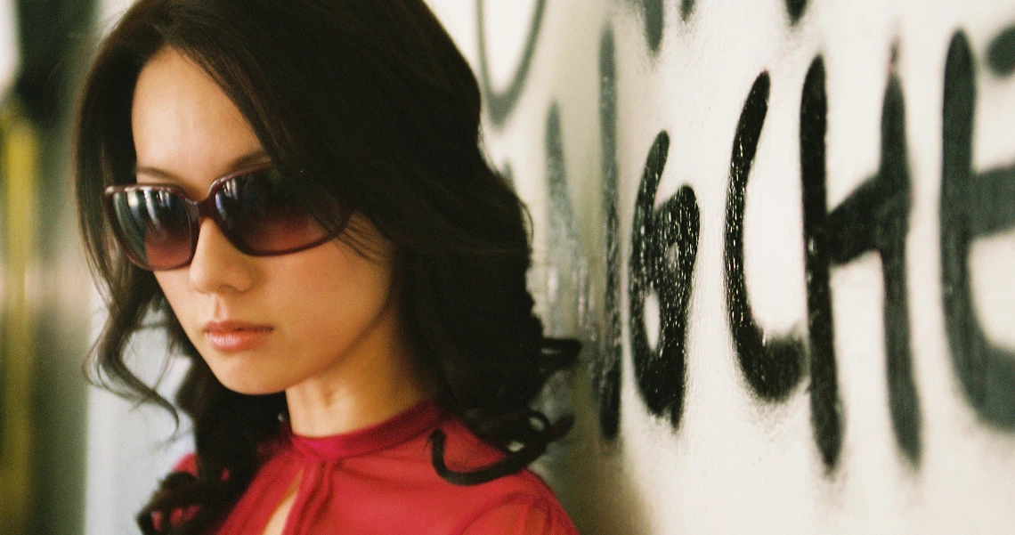 A woman with dark hair and sunglasses in a red top, from a film still of "Truth Be Told". Graffiti visible in background.