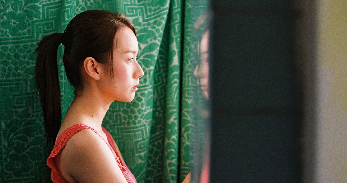 A woman in a red top gazes pensively out a window in a still from the film "Truth Be Told", showcasing introspective mood.