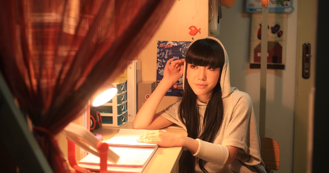 A film still from "Turn You Around" showing a young woman studying at a desk with warm lighting and red curtains.