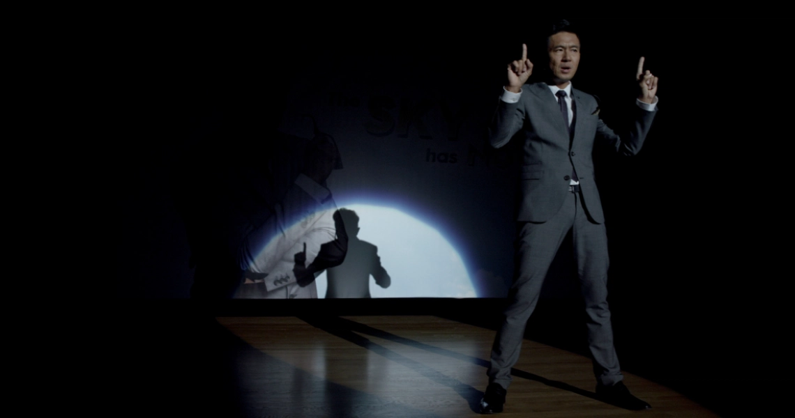 A scene from the film Unlucky Plaza showing a man in a suit gesturing dramatically, with a shadowy figure projected behind him.