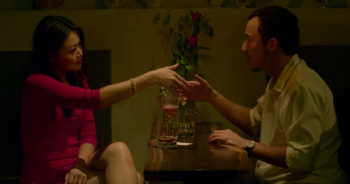 A tense scene from the film Unlucky Plaza shows a woman in pink handing a glass to a man in a dimly lit room.