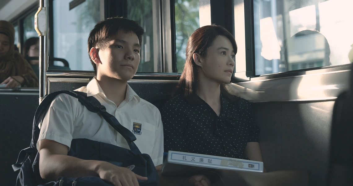 A scene from the film "Wet Season" showing two characters sitting side by side on public transport, looking pensive.