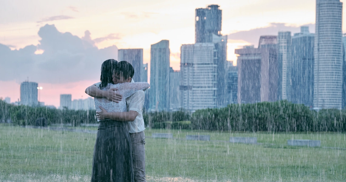 A couple embraces in the rain with Singapore's skyline in the background, a poignant scene from the film "Wet Season".