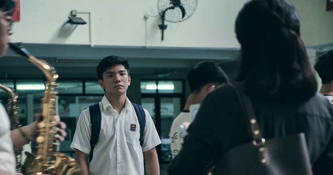 A student stands in a school hallway surrounded by classmates with musical instruments in a scene from the film "Wet Season".