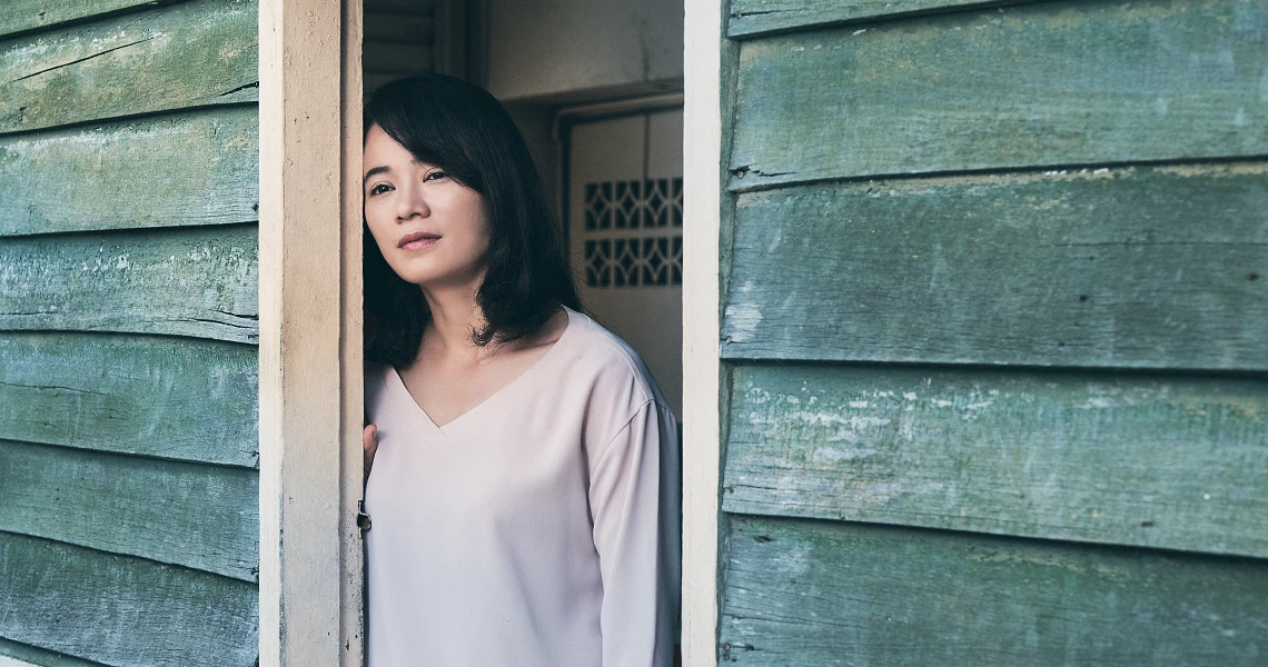 A woman in a white sweater stands in the doorway of a weathered green building in a scene from the film "Wet Season".