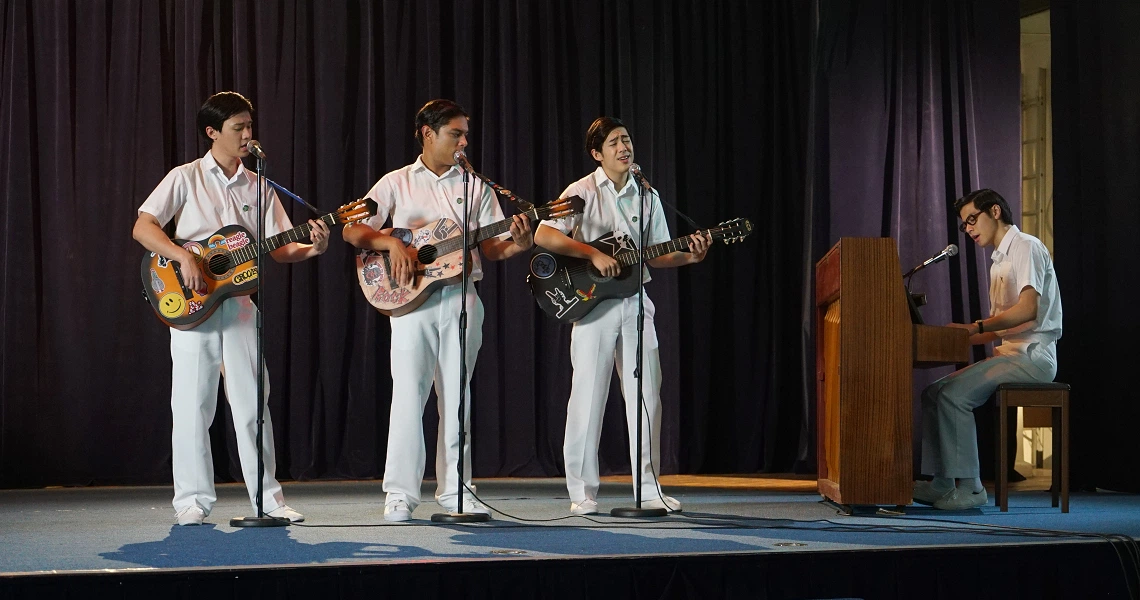 Four young men in white uniforms perform on stage in a scene from the film Wonder Boy, with three playing guitars and one at a piano.