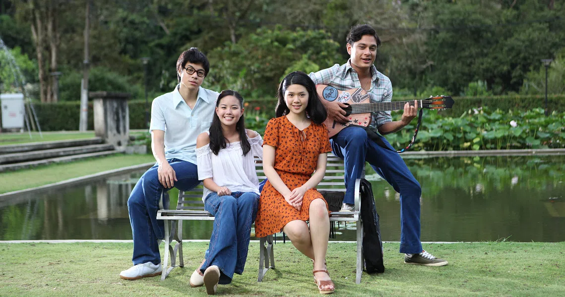 Four young actors from the film Wonder Boy sit on a bench near a pond, with one playing a guitar in an outdoor setting.