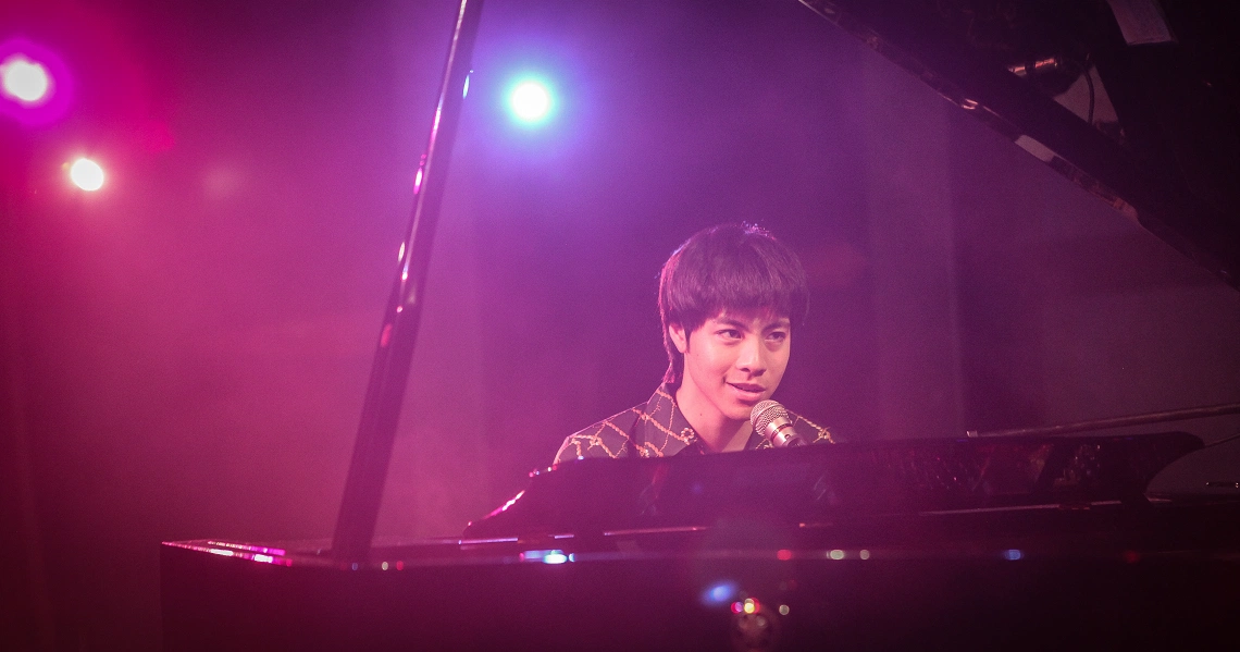 A young man plays piano on stage in a scene from the film Wonder Boy, bathed in vibrant purple and pink stage lighting.