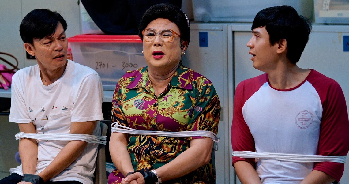 Liang Xi Mei, a middle-aged woman in colorful attire, sits between two young men in the film "Wonderful Liang Xi Mei".