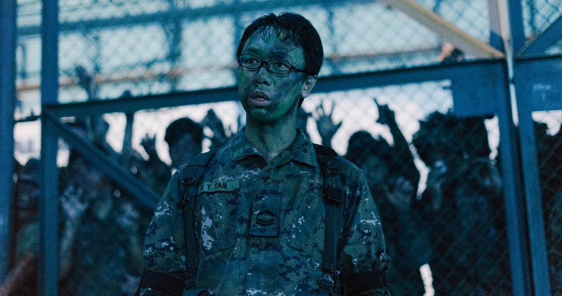 A soldier with blue-tinted skin stands in the foreground of a scene from the film Zombiepura, with shadowy figures visible behind a fence.